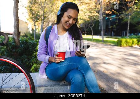 Donna asiatica sorridente che indossa le cuffie utilizzando lo smartphone e tiene il caffè da asporto in un parco soleggiato Foto Stock