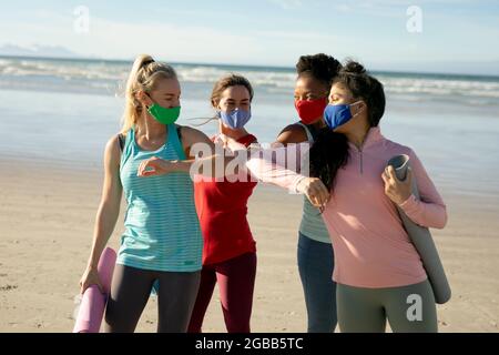 Gruppo di diverse amiche che indossano la maschera facciale che pratica yoga, in piedi e in spiaggia Foto Stock