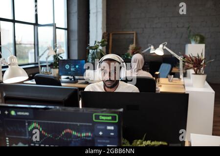 Giovane uomo d'affari africano in cuffia che lavora di fronte al monitor del computer in ufficio Foto Stock