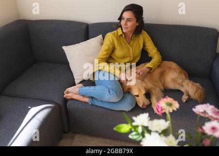 Donna caucasica che indossa una camicia gialla e si siede sul divano con il cane Foto Stock