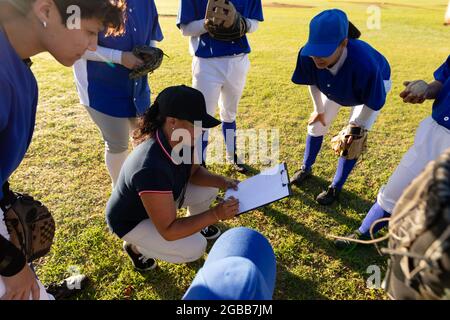 Gruppo vario di giocatori di baseball femminile in huddle intorno squatting allenatore femminile con appunti Foto Stock