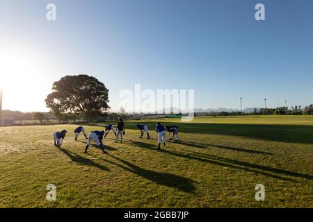 Gruppo variegato di giocatori di baseball femminile con allenatore che si riscalda in campo soleggiato, stretching Foto Stock