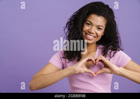 Giovane donna nera sorridente e mostrante il gesto del cuore isolato su sfondo viola Foto Stock