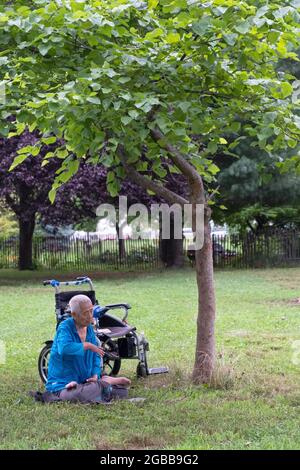 Un uomo seduto di 86 anni fa Falun Gong slow-moving esercizi vicino ad un albero. In un parco a Queens, New York, un luogo molto diverso. Foto Stock