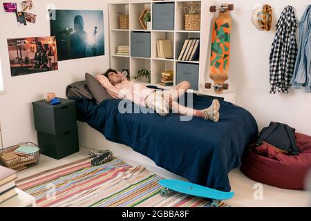 Giovane uomo sdraiato a letto tra scaffali con libri e skateboard mentre si riposa e pensa Foto Stock