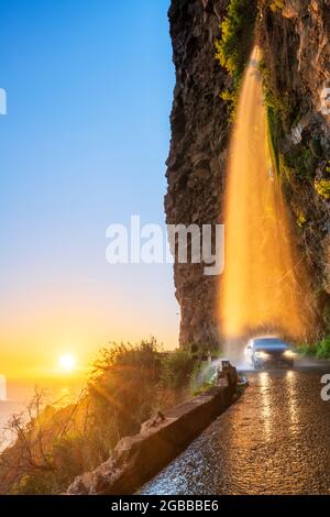 Auto che passa sotto la cascata di Anjos su strada costiera scivolosa al tramonto, Ponta do Sol, isola di Madeira, Portogallo, Atlantico, Europa Foto Stock