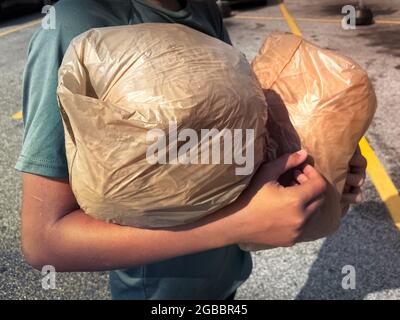 Braccia di giovane uomo che tengono i sacchetti di plastica della vendita al dettaglio impacchettati insieme per il riciclaggio mentre attraversano il parcheggio del supermercato. Foto Stock