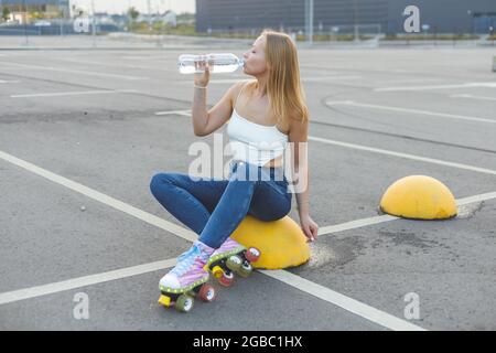 ragazza in skate-rollers seduta sul parcheggio e bere acqua dalla bottiglia. Foto Stock