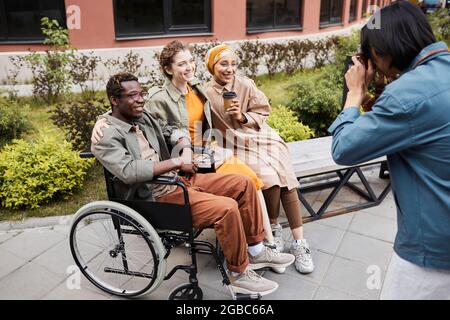 Uomo che fotografa un gruppo di amici multietnico, incluso un uomo disabile in carrozzina all'aperto Foto Stock