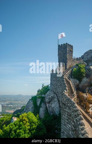 Dalle mura di un antico castello si vedono cittadine molto lontane ai piedi della montagna, un'antica bandiera portoghese sta lusinghando nel vento Foto Stock