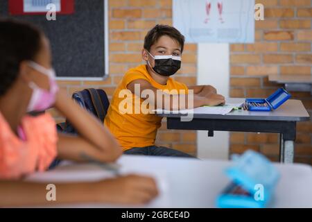 Ragazzo caucasico che indossa una maschera seduta sulla sua scrivania in classe a scuola elementare Foto Stock