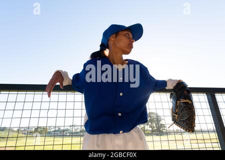 Giocatore misto di baseball femminile che indossa un guanto, appoggiandosi sulla recinzione al sole prima della partita Foto Stock