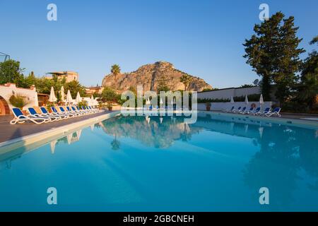 Cefalù, Palermo, Sicilia, Italia. Le torreggianti scogliere illuminate dal sole di la Rocca si riflettono nella tranquilla piscina dell'Hotel Kalura, alba. Foto Stock