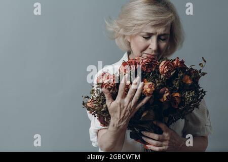 Triste donna anziana che tocca bouquet di fiori di rosa appassiti. Donna matura con capelli grigi in depressione. Concetto di invecchiamento vecchio. Riprese in studio isolate su sfondo grigio. Spazio di copia Foto Stock