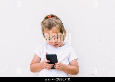 Bambina bionda che indossa una t-shirt bianca, guardando il telefono cellulare, con viso concentrato Foto Stock