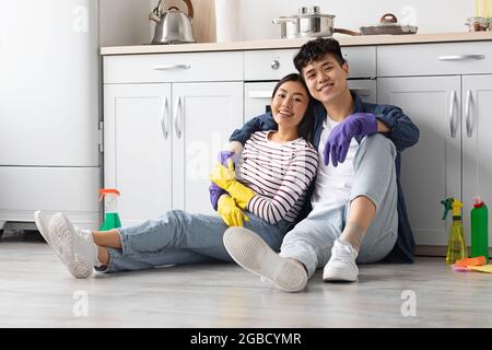 Coppia asiatica positiva seduta sul pavimento al kicthen, abbracciando Foto Stock