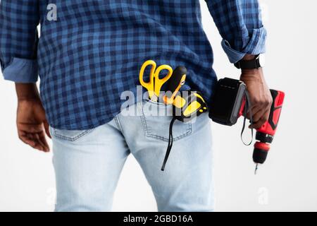 Utensile per foratura e fissaggio. Uomo afroamericano che tiene il trapano elettrico, il nastro di misurazione e le forbici in tasca Foto Stock