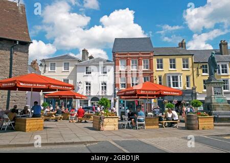 Visitatori persone seduti sotto Welcome Brecon ombrelloni a tavoli da picnic fuori della chiesa di St Marys nel centro città estate 2021 Powys Wales UK KATHY DEWITT Foto Stock