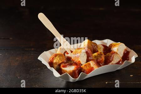 Appetitosi pezzi di salsiccia di maiale fritta con ketchup al curry e bastone ecologico in contenitore per andare su sfondo scuro Foto Stock