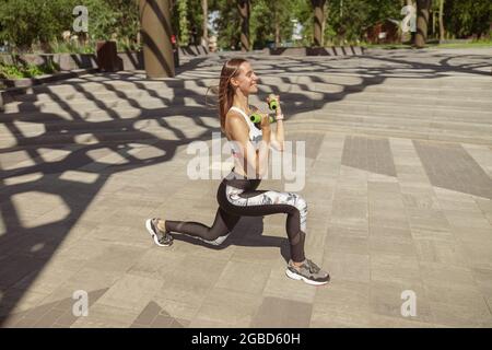 Bella donna sportiva in tuta fa affondo dinamico con manubri nel parco Foto Stock