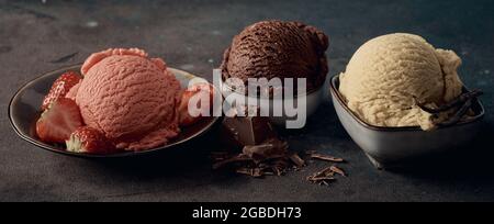 Ciotole con deliziosi cucchiai di cioccolato alla fragola e gelato alla vaniglia su un tavolo scuro Foto Stock