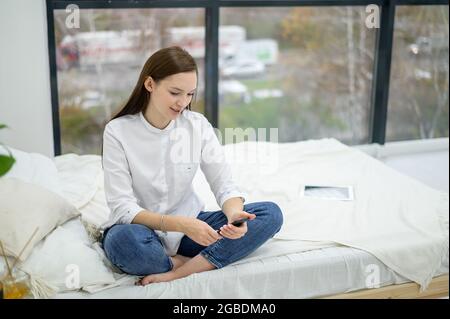 Una giovane brunetta in una camicia bianca e jeans scrive SMS sul telefono. Una ragazza si siede sul letto e legge il testo del messaggio sullo smartphone. Camera d'albergo Foto Stock