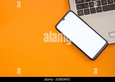 Smartphone con schermo bianco vuoto sulla tastiera del notebook. Telefono mockup. Vista dall'alto. Disposizione piatta. Isolato su sfondo arancione. Foto Stock