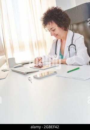 Una dottoressa usa il suo tablet digitale seduto alla scrivania del suo ufficio medico Foto Stock