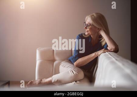 Felice donna matura in occhiali sorridente mentre si rilassa su un divano a casa nel soggiorno Foto Stock