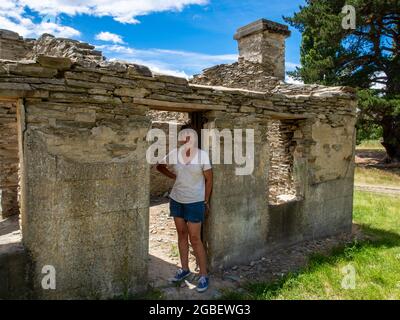 Una donna turistica esplora i resti di un vecchio hotel e di una cabina in un campeggio pubblico vicino al Passo Lindis nel centro di Otago, Nuova Zelanda Foto Stock