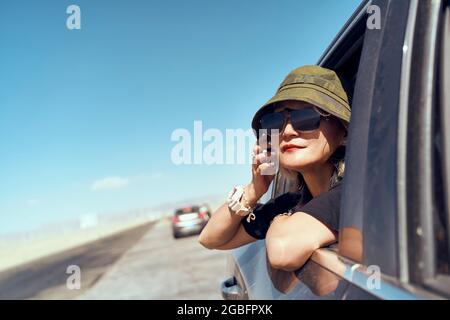 donna asiatica viaggiatore con cappello e occhiali da sole attaccando testa fuori dal finestrino posteriore dell'auto guardando alla vista Foto Stock