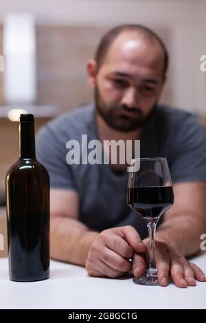 Primo piano di bottiglia e bicchiere di vino preparato per l'uomo che si sente infelice e solitario. Persona che beve bevande alcoliche, booze, liquore mentre è da solo in cucina. Adulto intossicato Foto Stock