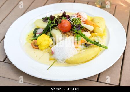 Verdure con pesce fritto e patate in salsa di zafferano, guarnite con erbe e schiuma su un piatto bianco e un tavolo di legno, fuoco selezionato, stretto de Foto Stock