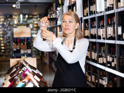 Produttore di vino donna matura e focalizzato che ispeziona la qualità del vino in cantina sullo sfondo con ripiani di bottiglie di vino Foto Stock