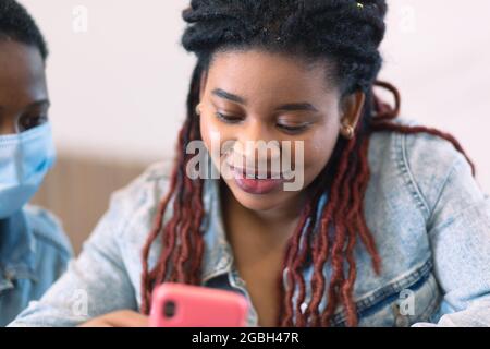 Una donna afroamericana si siede con un telefono rosa nelle mani. Un'altra donna coetanei sopra la sua spalla. Foto Stock