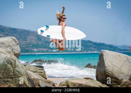 Donna sorridente che salta in aria con la sua tavola da surf, spiaggia di Punta Paloma, Tarifa, Cadice, Andalusia, Spagna Foto Stock