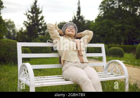 Felice delizia anziana donna con capelli grigi che guarda in su seduta sulla panchina nel parco Foto Stock