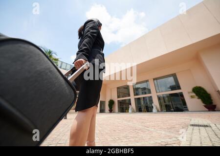 Sotto la vista di una donna d'affari in formalwear nero che tiene la maniglia della valigia gommata e che guarda all'entrata dell'edificio dell'aeroporto Foto Stock