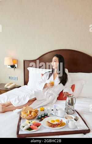 Giovane donna asiatica in accappatoio sdraiato a letto e bere succo mentre si serve la colazione in hotel Foto Stock
