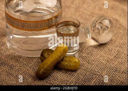 Un decanter di vetro con vodka, un bicchiere di vodka e cetrioli decapati su uno sfondo di tessuto casalinghi con una consistenza ruvida. Primo piano fuoco selettivo Foto Stock