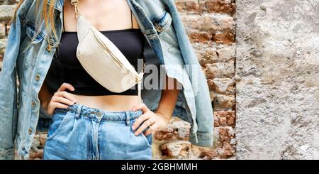 Giovane bella donna tagliata in piedi accanto al muro di mattoni di strada, jeans jeans jeans vestiti, cintura di tenuta borsa a mano in spalla, tendenza moda estate, Foto Stock