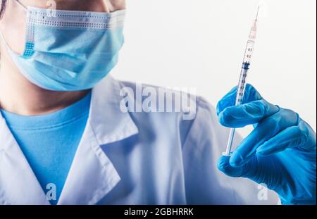 Ago di closeup del vaccino covid-19 in mano a uno scienziato professionista o medico in laboratorio per il trattamento con guanti da maschera e camice da laboratorio sulla parte posteriore bianca Foto Stock