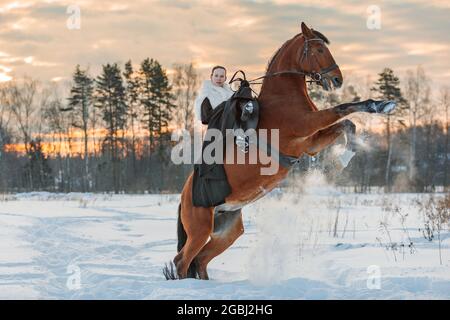 Una ragazza in un mantello bianco cavalca un cavallo marrone in inverno. Ora d'oro, sole tramontante. Il cavallo si alza. Mosca, Russia 22 febbraio 2021 Foto Stock