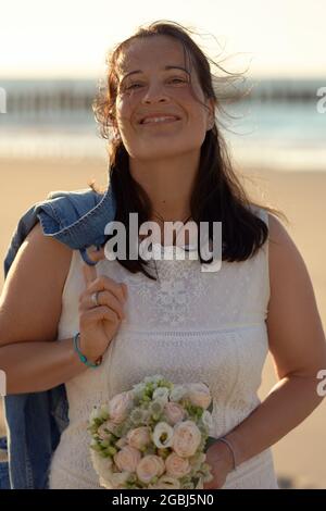 Donna moderna e felice in un elegante abito bianco con una giacca in denim che si è schiantata sopra la spalla sorridendo alla macchina fotografica mentre porta un bouque nuziale Foto Stock