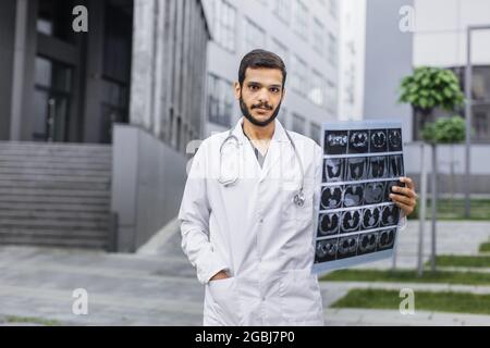 Vita in su ritratto esterno di giovane bel medico indiano arabo sicuro maschile, guardando la fotocamera, tenendo la tomografia del paziente scansione, in posa vicino all'ospedale moderno. Concetto di radiologia Foto Stock