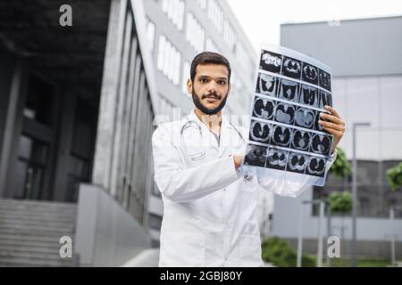 Vita in su ritratto esterno di giovane bel medico indiano arabo sicuro maschile, guardando la fotocamera, tenendo la tomografia del paziente scansione, in posa vicino all'ospedale moderno. Concetto di radiologia Foto Stock