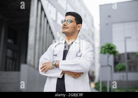 Primo piano ritratto di un giovane medico arabo maschile professionista, indossando cappotto bianco e stetoscopio intorno al collo, guardando da sogno, in piedi all'aperto di fronte al moderno edificio dell'ospedale Foto Stock