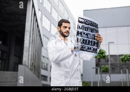 Radiologo arabo specializzato in medicina maschile di alto livello in camice bianco da laboratorio, che guarda i raggi X rm dei polmoni del paziente, in piedi all'esterno di fronte alla clinica moderna Foto Stock
