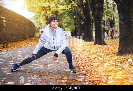 Donna atletica in forma che fa allenamento prima di fare jogging nel parco cittadino autunnale sul parco giochi per bambini. Giovane fitness femminile runner che allunga le gambe mentre warmin Foto Stock