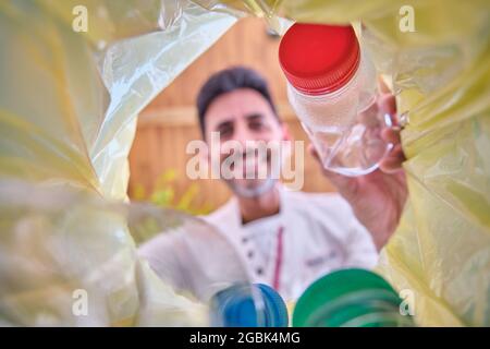 immagine dall'interno di un cestino di riciclaggio con un sacchetto giallo di un uomo che lancia una bottiglia di plastica con un tappo rosso per riciclare in cui il suo viso è fuori fuoco Foto Stock